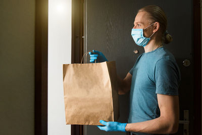 Man holding paper bag with food in doors, in medical gloves and protective mask, copy space. safe