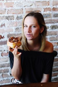 Close-up portrait of young woman eating dessert while sitting against brick wall