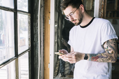 Confident male hipster hacker text messaging through smart phone by window at creative office