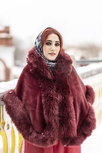 A slavic woman in a national colored scarf,a fur coat and  mittens on the porch of a wooden house