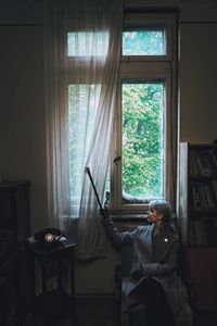 Woman looking through window while sitting on chair at home