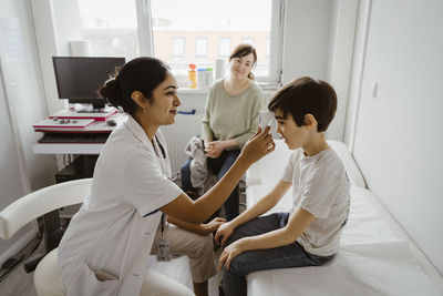 Smiling female pediatrician measuring body temperature of boy through infrared thermometer at clinic