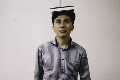 Portrait of man with book on head while standing against white wall