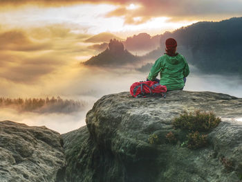 Alone girl sits with her back on a cliff against huge misty mountain