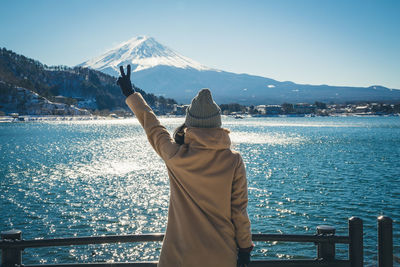 Rear view of woman gesturing peace sign while standing against lake in winter