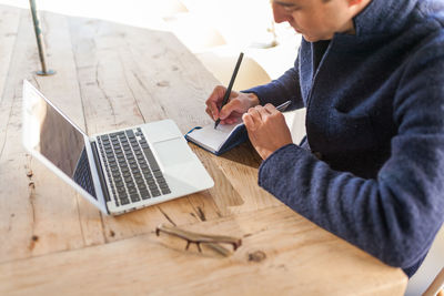 Side view of concentrated young bearded male freelancer in casual clothes and eyeglasses sitting at table with laptop and smartphone while working on project in outdoor cafe