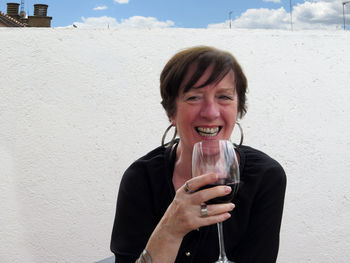 Portrait of smiling woman holding wineglass while sitting on chair against wall