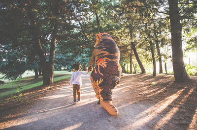 Rear view of boy walking with person wearing dinosaur costume in park