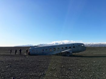 People by crashed airplane wreck on black sand against blue sky