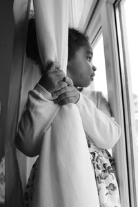 Low angle view of thoughtful girl holding curtain while looking through window at home