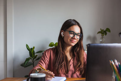 Happy young female in wireless earbuds and eyeglasses sitting at table with cup of hot drink and typing on modern netbook at home