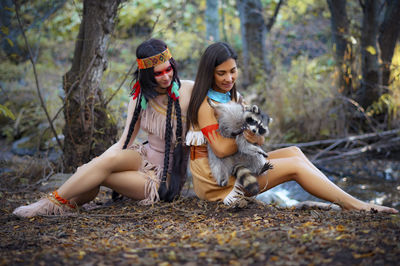 Young women in traditional clothing sitting with raccoon in forest