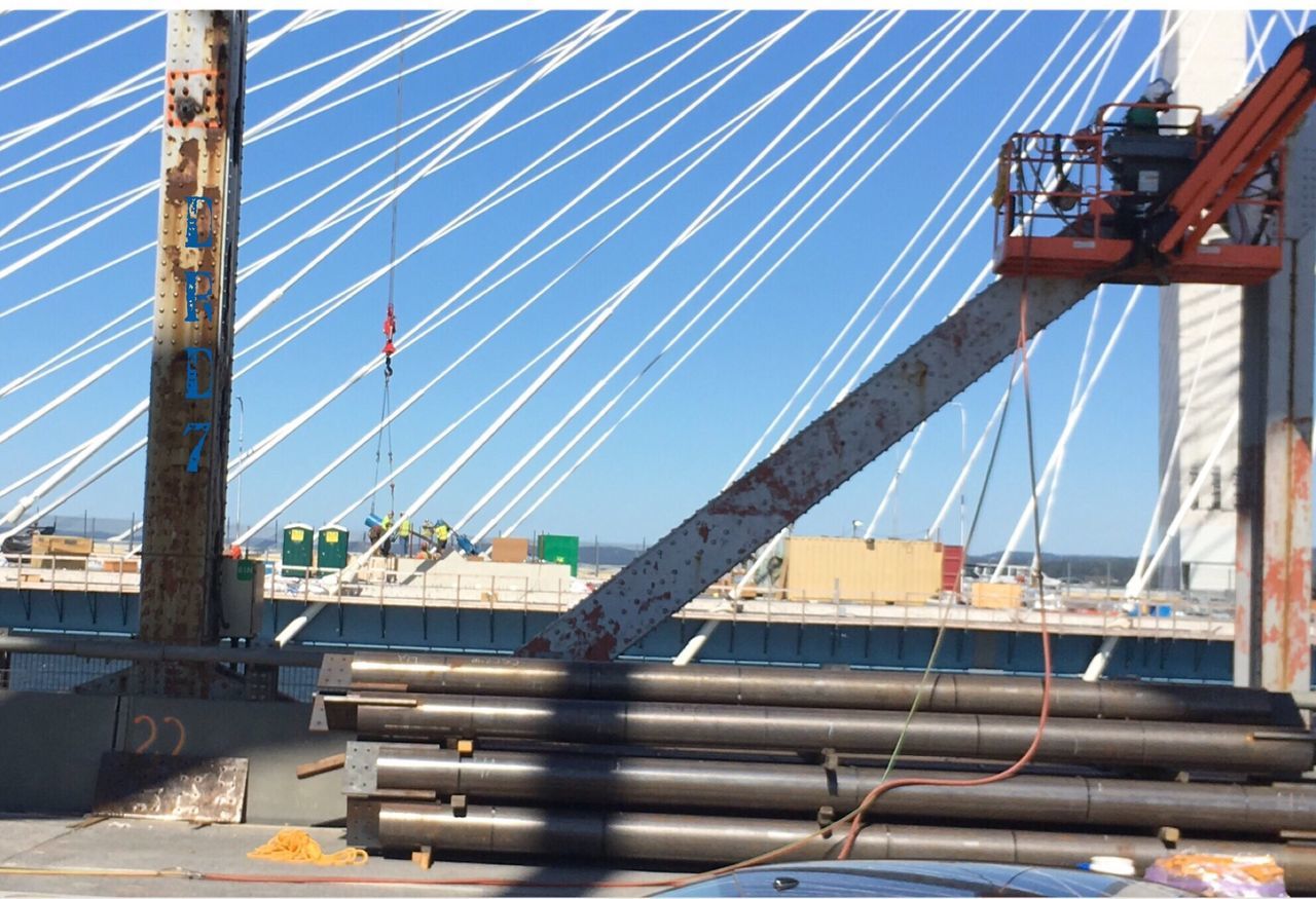 transportation, bridge - man made structure, connection, cable, built structure, mode of transport, day, architecture, outdoors, city, sunlight, no people, sky, industry, suspension bridge, girder, building exterior, nautical vessel, clear sky, harbor, water, shipyard