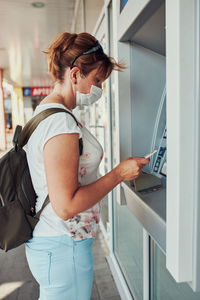 Woman withdrawing money from atm machine using debit or credit card standing outdoors in the street