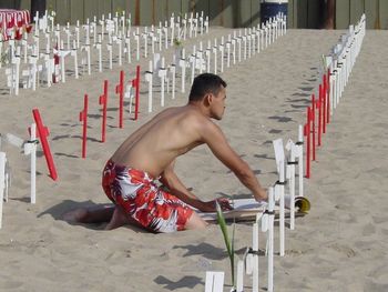 Shirtless man kneeling by cross on sand at beach