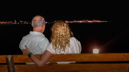 Rear view of mature couple sitting against sea on bench at night