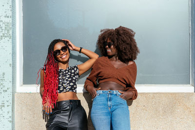 Black sisters in sunglasses standing on pavement and leaning on building wall while chilling in daylight