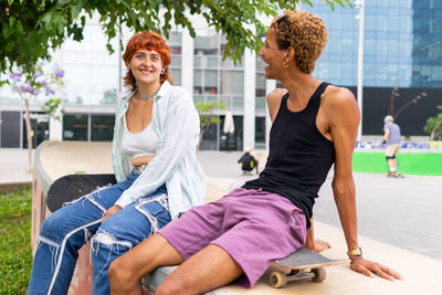 Positive multiracial friends in casual wear with skateboards sitting together on border near modern building in city on summer day