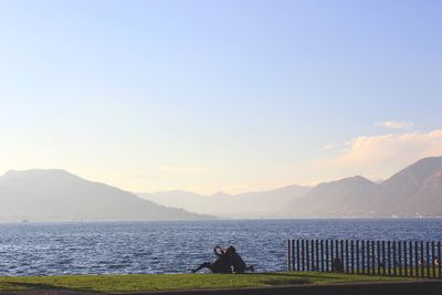 Rear view of men sitting on sea by mountains against sky