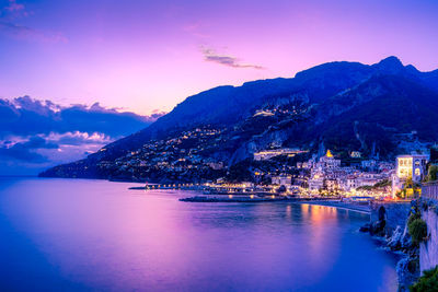 Illuminated city by sea against sky during sunset