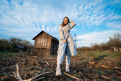 Beautiful girl with long hair in a grey trench coat next to an old wooden house blue sky background 