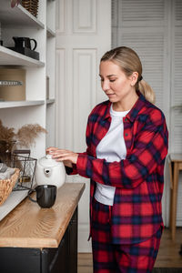 A millennial girl in homemade pajamas pours herself tea while standing in her cozy kitchen