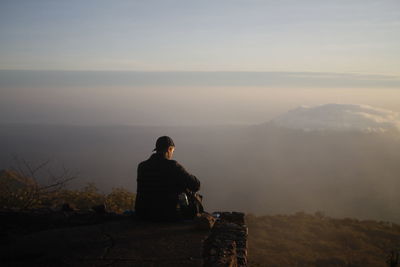 Rear view of man sitting on rock against sky