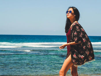 Young woman wearing sunglasses standing at beach against clear sky