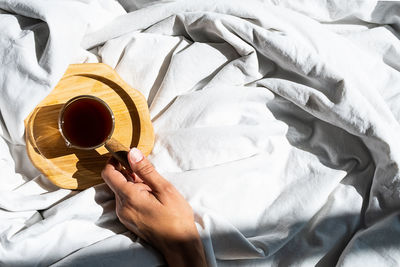 Females hand reaches for morning coffee cup in bed. woman wakes up on white bedding in sunlight.