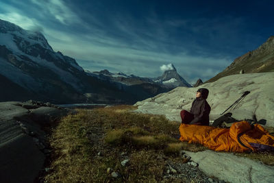 Mature woman sitting on field amidst mountains against cloudy sky