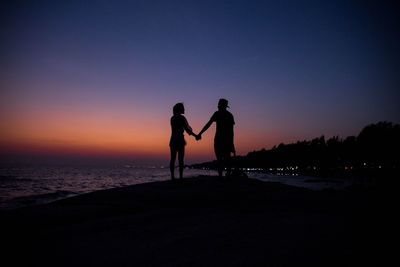 Silhouette of couple holding hands while standing at beach against sky during sunset
