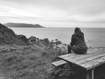 Rear view of woman sitting on bench at sea shore against sky