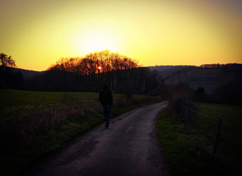 Rear view of woman on road amidst field against sky during sunset