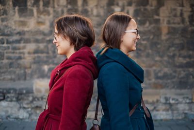 Smiling sisters standing back to back against stone wall