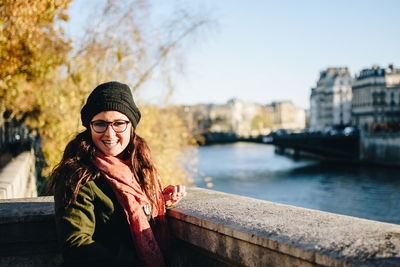 Portrait of smiling young woman by river in city against sky