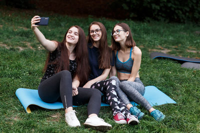Three girls sitting on yoga mat and doing selfies in green park
