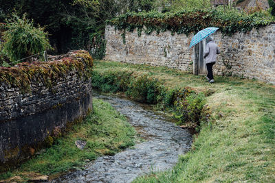 Rear view full length of senior man with umbrella walking by stream on grass