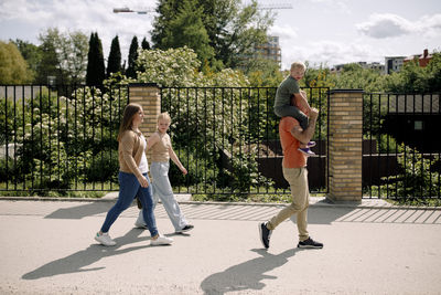 Father carrying son on shoulders while walking ahead of woman and daughter at park during sunny day