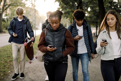 Multi-ethnic teenage friends using smart phones while walking on street in city