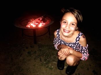 Portrait of smiling girl crouching by fire pit at night