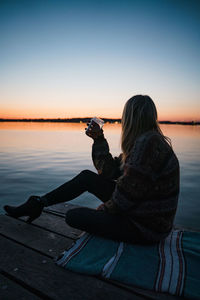 Woman sitting by sea against sky during sunset