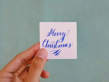 Close-up of hand holding paper with christmas text against wall