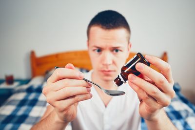 Sick man pouring medicine from bottle in spoon while sitting on bed at home