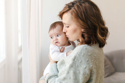 Loving mother kissing cute baby near window at home