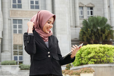 Young woman using smart phone while standing against buildings