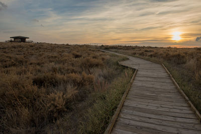 Boardwalk amidst field against sky during sunset