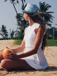 Beautiful woman holding coconut while sitting on beach