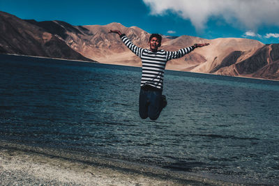 Full length of young man with arms raised jumping at lakeshore