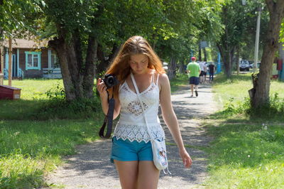 A red-haired girl walks with a camera along the alley at the campsite, on a sunny summer day.