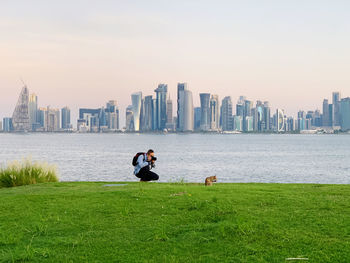 Man photographing animal on grassy land against river and modern buildings